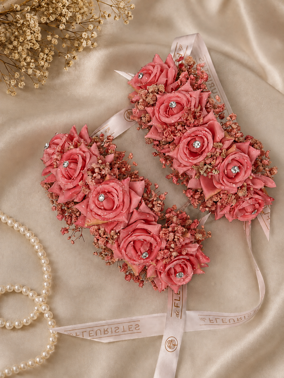 Two pink fabric garter straps decorated with artificial roses and baby's breath, tied with white ribbons printed with the brand name 'Zell & Feuren', displayed on a beige satin fabric with a pearl necklace and dried flowers as props.