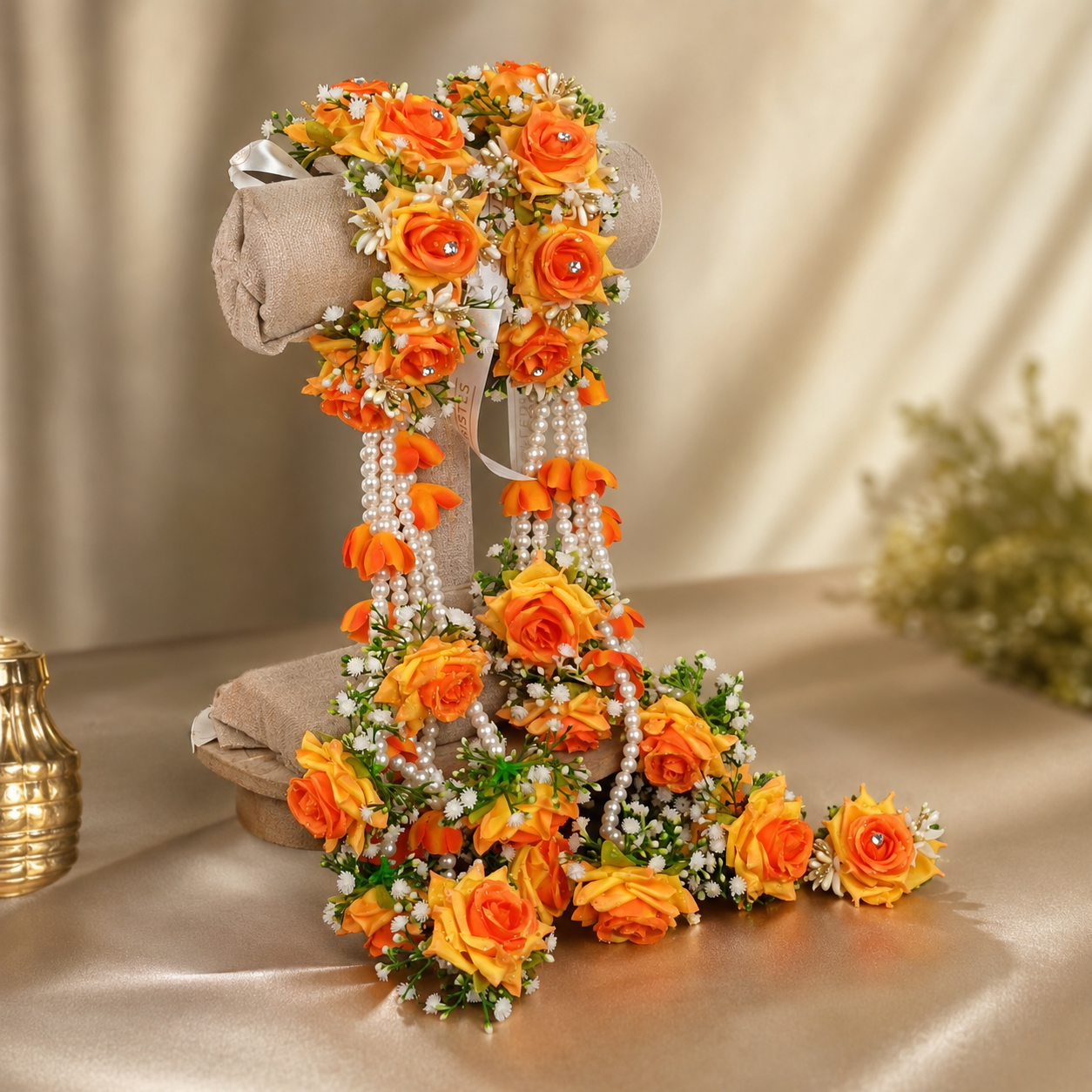 An elaborate floral garland arrangement displayed on a beige mannequin stand, featuring bright orange and yellow artificial roses, green leaves, and strands of white faux pearls, set against a draped beige fabric background.