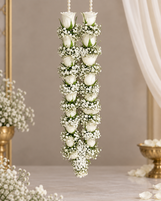 A hanging floral garland composed of white roses and clusters of small white baby's breath, suspended against a softly lit indoor backdrop with draped fabric and gold-toned decor in the background.
