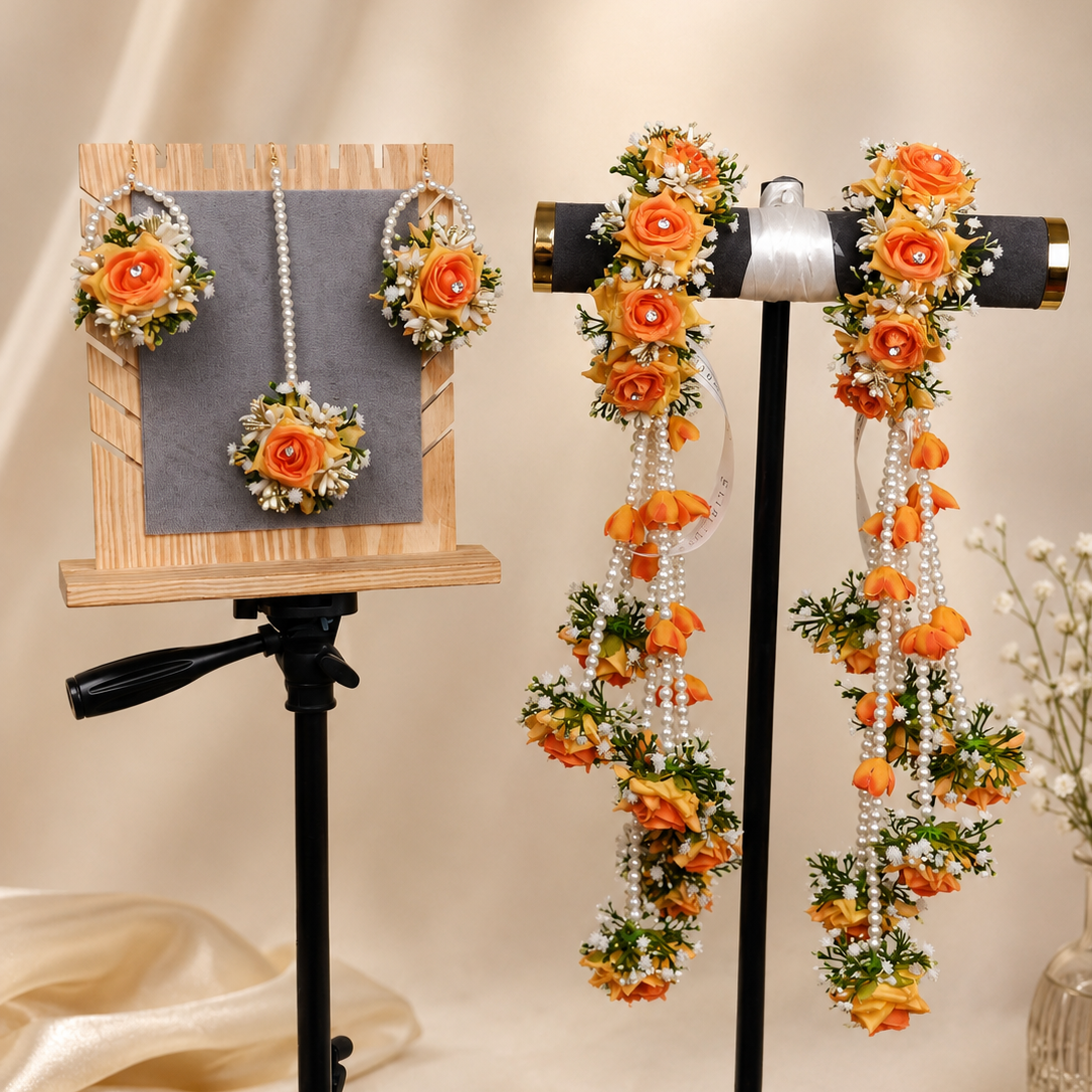 Orange and white floral garland jewelry accessories displayed on black stands against a beige draped fabric background. The set includes matching flower earrings on a wooden display board and long floral garlands with pearl strands on a horizontal stand.