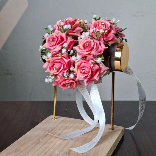 A pink artificial rose bouquet with small white filler flowers and green leaves, mounted on a stand with a gold cap and white ribbon, displayed on a wooden board against a gray wall.