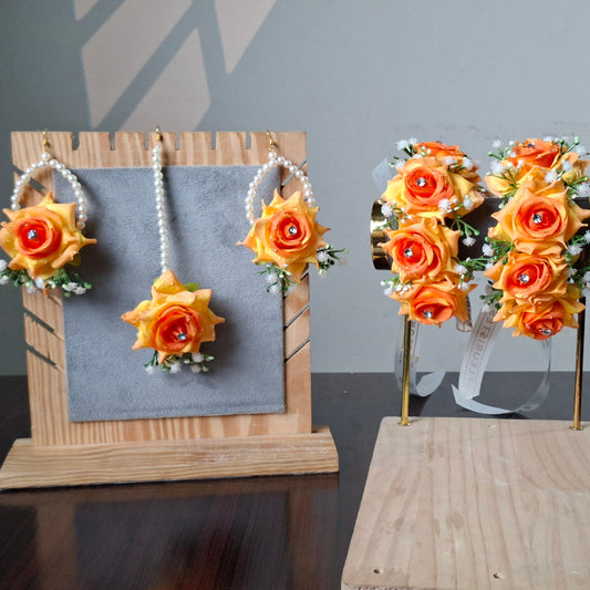 A set of artificial flower accessories displayed on stands, including two matching yellow and orange rose wristlets with pearl-like ribbons and two matching rose bachelorette necklaces, shown on a dark table against a gray wall.