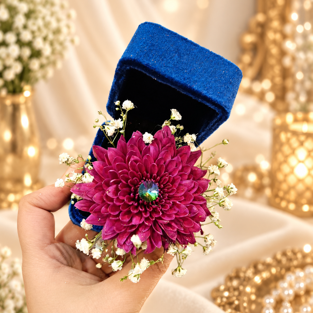 A hand holds an open royal blue velvet ring box decorated with a large magenta dahlia flower and small white baby's breath, set against a warm, softly lit background with gold decor and draped fabric.