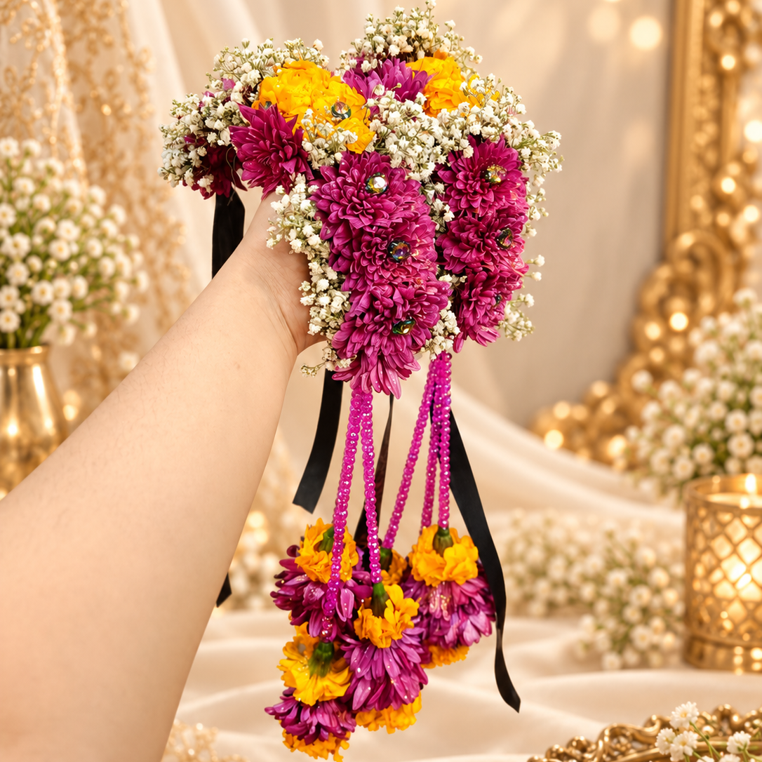 A hand holding a traditional Thai flower bouquet (khon) featuring vibrant magenta chrysanthemums, bright yellow marigolds, and delicate white baby's breath, with hanging black ribbons and purple bead garlands, set against a warm, decorated background of gold mirrors and draped white fabric.