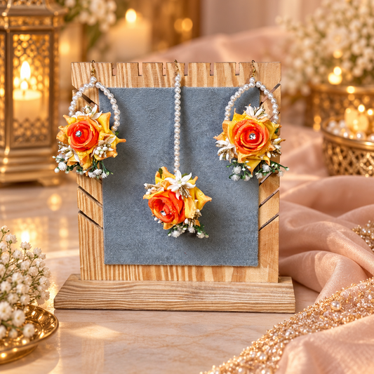 A jewelry display stand holding a matching floral earring set. The set includes two drop earrings and one pendant, decorated with orange and yellow artificial roses, white pearl strands, and small white flowers with green leaves.