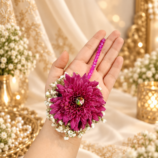 A close-up of a hand holding a magenta floral wrist accessory with a matching magenta beaded strap, set against a warm, decorated background with gold and cream fabrics.