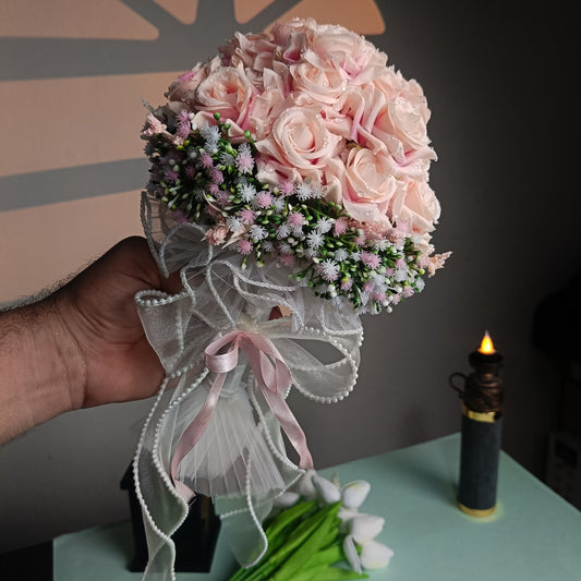 A hand holds a bouquet of light pink roses with green and purple filler flowers, tied with a sheer white ribbon and bow. The bouquet is photographed indoors on a table with a candle and tulips in the background.